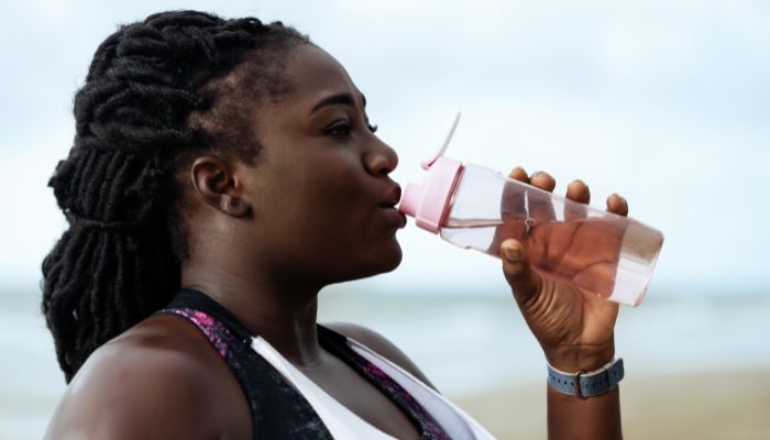 Imagem de mulher negra bebendo água na praia. Ela usa um squeeze transparente.