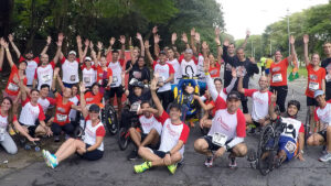Imagem de voluntários do Pernas de Aluguel levantam as mãos reunidos com pessoas com deficiências em cadeiras de rodas.