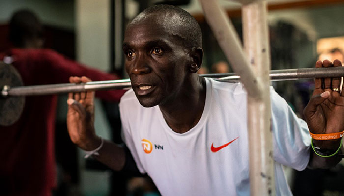 Imagem de Eliud Kipchoge fazendo fortalecimento muscular na academia. Ele está com uma barra nas costas, fazendo agachamento.