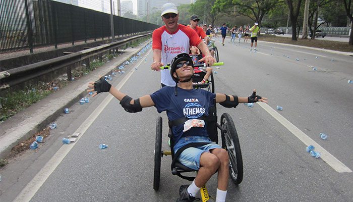 Imagem de voluntário levando um jovem em cadeira de rodas. O jovem deficiente está de braços abertos e sorrindo.