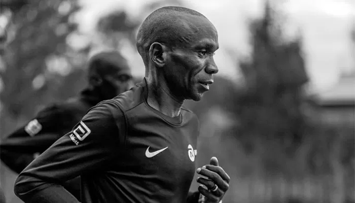 Imagem em preto de branco de Eliud Kipchoge de perfil enquanto corre ao lado de outro atleta.