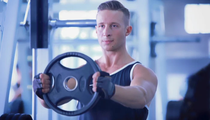 Homem vestindo regata azul está no centro da imagem levantando uma anilha em uma academia.